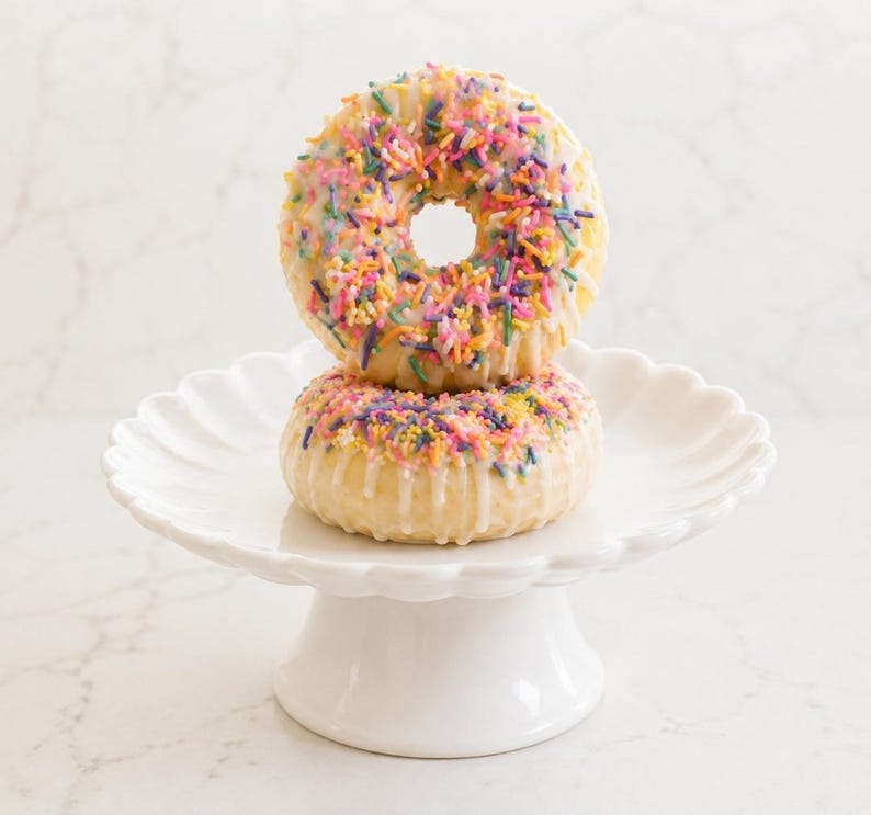 May include: Two stacked ring-shaped pastries with white icing and colorful sprinkles on a white scalloped pedestal stand. The top pastry is heavily covered in pink, yellow, blue, and purple sprinkles, while the bottom pastry has fewer.