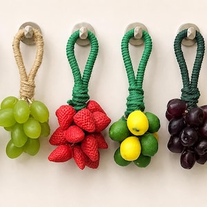 May include: Six decorative fruit ornaments hanging from silver wall hooks. Each ornament features a cluster of artificial fruit, including green limes, green grapes, red strawberries, mixed limes and lemons, dark purple grapes, and yellow lemons.