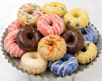 May include: A collection of assorted glazed donuts arranged on a scalloped-edge metal pedestal stand. The donuts feature various toppings including colorful sprinkles, white icing drizzles, and solid glazes in shades of pink, brown, blue, and yellow.