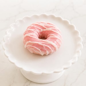 May include: A round donut with a smooth pink glaze and a white icing drizzle, centered on a white ceramic cake stand that features a scalloped edge design. The donut is positioned in the middle of the plate, showcasing the contrast between the pink.