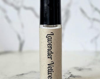 May include: A clear glass roller bottle with a black cap, featuring a tan paper label wrapped around the center. The label has the text Lavender Vetiver printed in a black, stylized serif font oriented vertically along the bottle body.