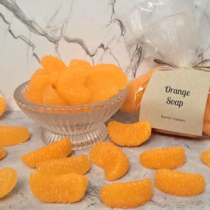 May include: A collection of bright orange, crescent-shaped soap segments resembling mandarin slices. Some segments are arranged in a clear glass bowl, while others are scattered around. A clear plastic bag contains more soap with a label reading Orange Soap.