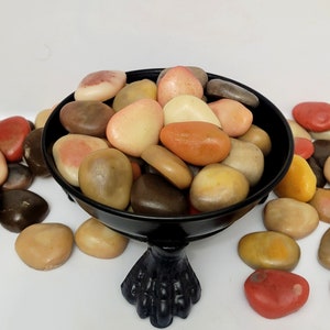 May include: A black bowl with a decorative foot sits on a white surface. The bowl is filled with smooth, colorful stones in shades of brown, orange, pink, and white.
