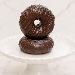 May include: Two round chocolate-covered donuts stacked vertically on a white scalloped pedestal stand. The top donut features a textured, drizzled chocolate glaze, while the bottom donut has a smooth, glossy dark brown chocolate coating.