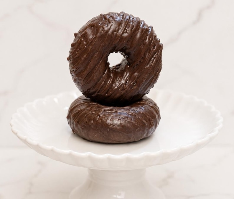 May include: Two round chocolate-covered donuts stacked vertically on a white scalloped pedestal stand. The top donut features a textured, drizzled chocolate glaze, while the bottom donut has a smooth, glossy dark brown chocolate coating.