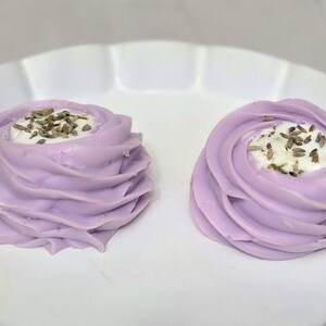 May include: Two lavender-colored, swirl-shaped pastries with a white filling and topped with seeds, presented on a white, ruffled-edge plate. The pastries have a soft, creamy appearance, suggesting a dessert.