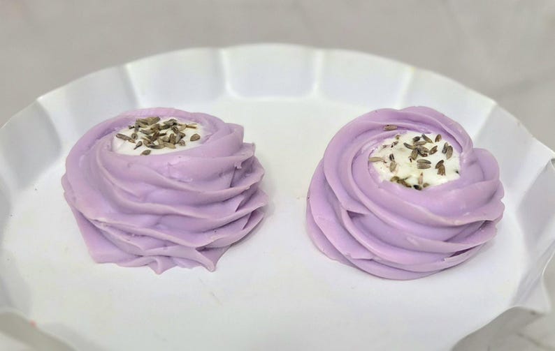 May include: Two lavender-colored, swirl-shaped pastries with a white filling and topped with seeds, presented on a white, ruffled-edge plate. The pastries have a soft, creamy appearance, suggesting a dessert.
