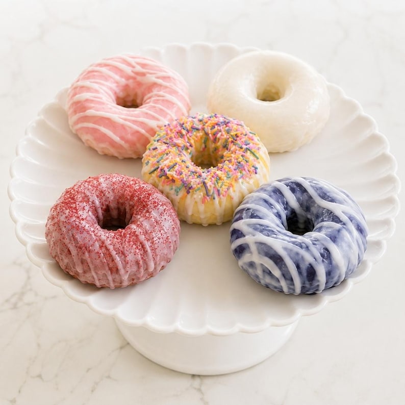 May include: Five glazed donuts arranged on a white scalloped cake stand. The donuts feature various toppings including pink icing with white drizzle, a plain white glaze, colorful sprinkles, red sugar crystals, and a purple glaze with white drizzle.