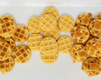 May include: A collection of small, round, golden-brown waffle-shaped items. Some pieces feature a grid pattern, while others are topped with a small, square, off-white pat of butter. The items are arranged in a loose cluster on a white surface.