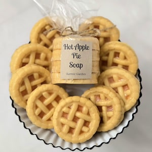 May include: A stack of round, yellow soap bars shaped like miniature lattice-top apple pies, wrapped in clear plastic with a tan paper label that reads Hot Apple Pie Soap and Karmic Garden, tied with a thin brown twine string at the top.
