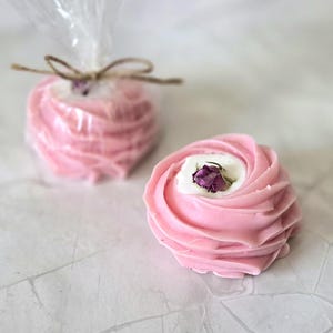 May include: Two pink, rose-shaped soaps with a white center and a dried rosebud. One soap is unwrapped, the other is wrapped in clear cellophane and tied with twine. The soaps are on a white surface.