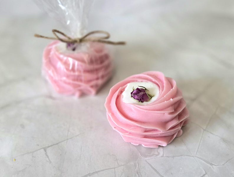 May include: Two pink, rose-shaped soaps with a white center and a dried rosebud. One soap is unwrapped, the other is wrapped in clear cellophane and tied with twine. The soaps are on a white surface.