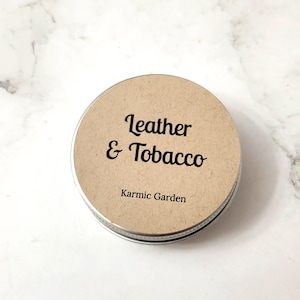 May include: A round metal tin with a brown paper label. The label says "Leather & Tobacco" and "Karmic Garden".