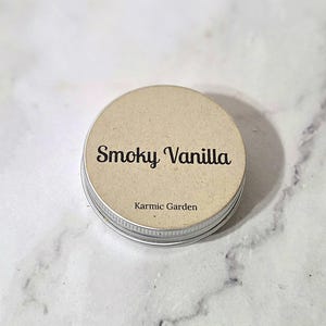 May include: A small, round metal tin with a tan paper label. The label reads "Smoky Vanilla" in a cursive font, with "Karmic Garden" below. The tin has a silver rim and a neutral color palette.