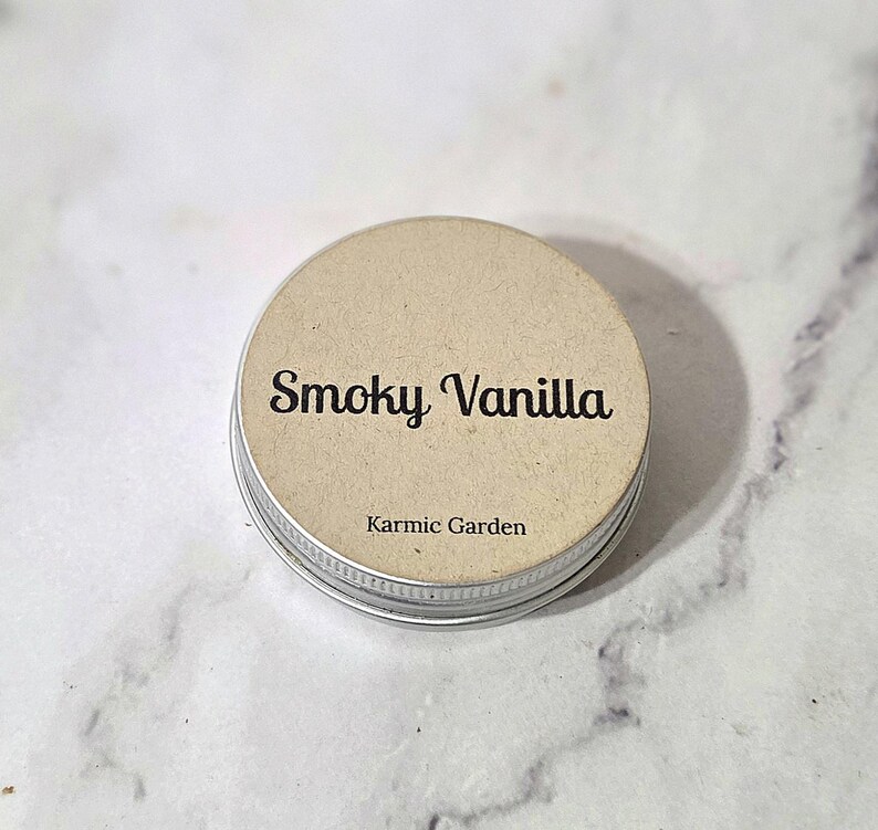 May include: A small, round metal tin with a tan paper label. The label reads "Smoky Vanilla" in a cursive font, with "Karmic Garden" below. The tin has a silver rim and a neutral color palette.