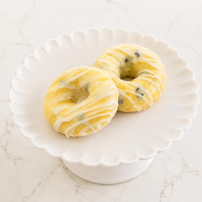 May include: Two round, golden-yellow blueberry donuts topped with a white icing drizzle, resting on a white ceramic pedestal cake stand with a scalloped edge. The donuts feature visible dark blue berries embedded within the light yellow dough.