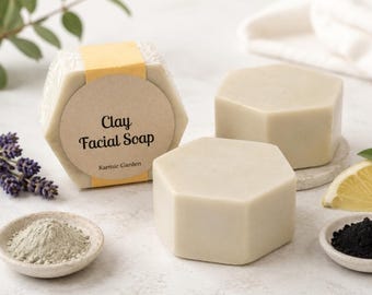 May include: Three hexagonal bars of light beige clay facial soap. One bar features a yellow paper band with a circular label reading Clay Facial Soap and Karmic Garden. Small bowls containing grey clay powder and black charcoal powder are nearby.