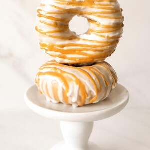 May include: Two round donuts stacked vertically on a small white pedestal stand. Each donut is coated in a smooth white glaze and decorated with thin, horizontal drizzles of golden-orange icing that wrap around the surface of the pastry.