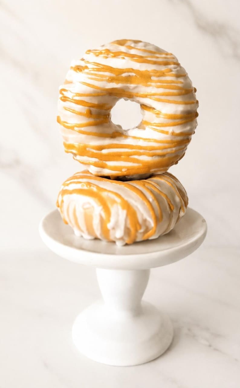 May include: Two round donuts stacked vertically on a small white pedestal stand. Each donut is coated in a smooth white glaze and decorated with thin, horizontal drizzles of golden-orange icing that wrap around the surface of the pastry.