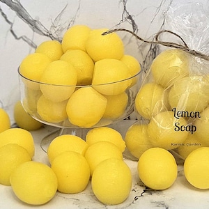 May include: A collection of small, round, bright yellow lemon-shaped soaps. Some soaps are piled in a clear glass pedestal bowl, while others are contained in a clear plastic bag tied with twine, labeled with the text Lemon Soap Karmic Garden.