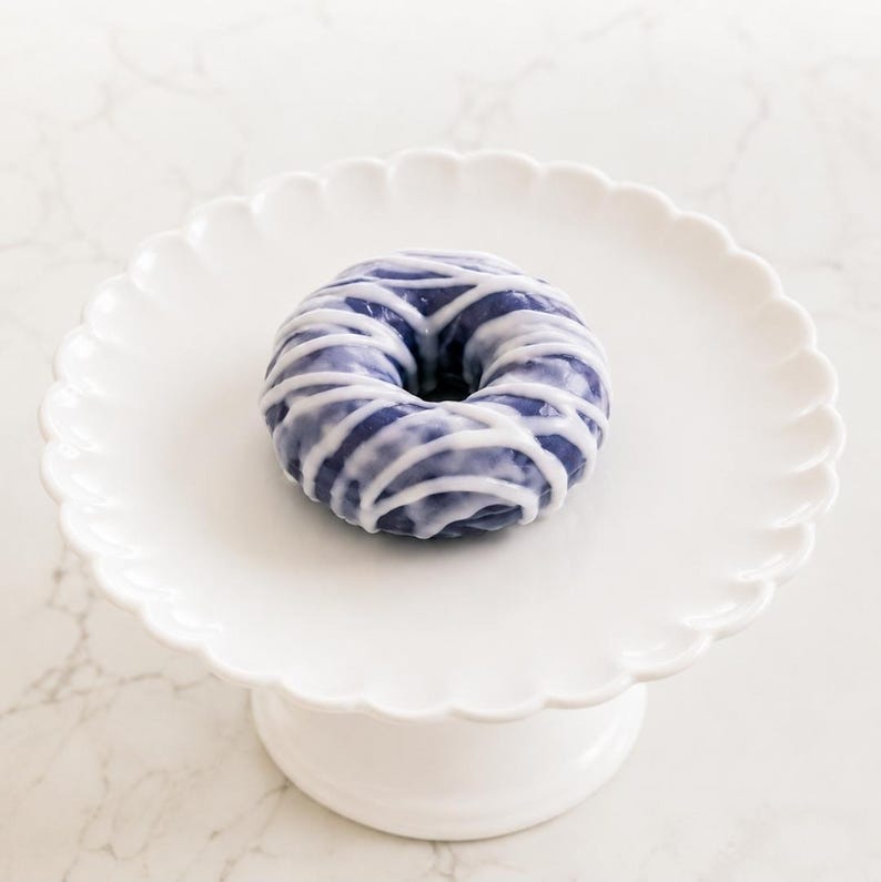 May include: A purple glazed donut with a white icing drizzle sits centered on a white scalloped cake stand. The donut features a smooth, deep violet surface texture contrasted by thin, intersecting lines of opaque white sugar glaze.
