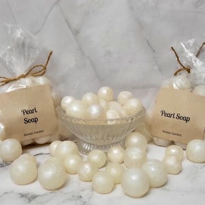 May include: Pearl soap in a clear glass bowl and in clear cellophane bags tied with twine. The bags have a brown paper label that reads "Pearl Soap" and "Karmic Garden". The soap is white and pearl-like.
