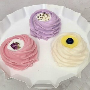 May include: Three decorative soaps in a white, ruffled dish. The soaps are swirled in shades of pink, lavender, and cream, each with a unique topping: a dried rose, lavender buds, and a blueberry.