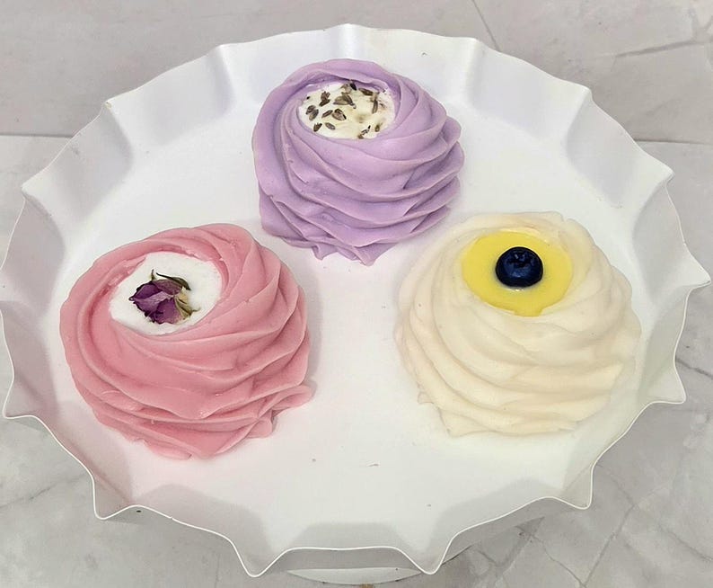 May include: Three decorative soaps in a white, ruffled dish. The soaps are swirled in shades of pink, lavender, and cream, each with a unique topping: a dried rose, lavender buds, and a blueberry.