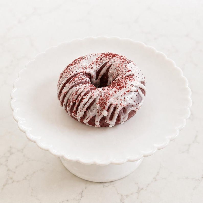 May include: A red velvet donut topped with a white icing drizzle and fine red sprinkles, centered on a white scalloped cake stand. The donut has a circular shape with a hole in the middle, showcasing a deep red color beneath the white glaze.