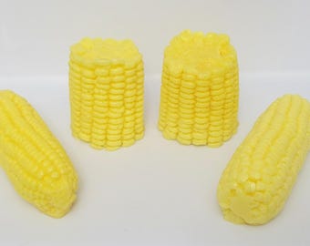 May include: Four bright yellow corn-shaped objects arranged on a plain white surface. Two pieces are upright, cylindrical segments of corn on the cob, while the other two are longer, slightly curved, full-length corn cobs with textured kernels.