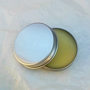 May include: A small, round, silver-colored tin with a screw-on lid. The lid is partially open, revealing a light green, balm-like substance inside. The tin sits on a white, textured surface.