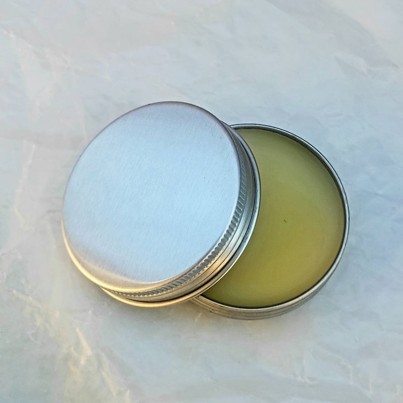 May include: A small, round, silver-colored tin with a screw-on lid. The lid is partially open, revealing a light green, balm-like substance inside. The tin sits on a white, textured surface.