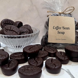 May include: Dark brown soap pieces shaped like coffee beans, some contained in a clear glass bowl and others scattered around. A clear plastic bag holds more soap pieces, featuring a tan label with the text Coffee Bean Soap and Karmic Garden.