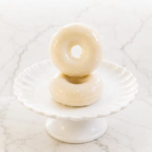 May include: Two glazed donuts stacked vertically on a white ceramic pedestal stand with a scalloped edge. The donuts have a smooth, shiny, off-white sugar glaze coating their entire surface, and the stand features a round base and a fluted rim.