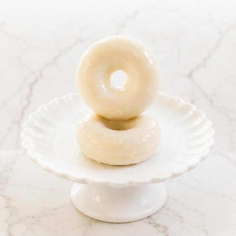 May include: Two glazed donuts stacked vertically on a white ceramic pedestal stand with a scalloped edge. The donuts have a smooth, shiny, off-white sugar glaze coating their entire surface, and the stand features a round base and a fluted rim.