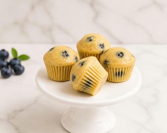 May include: Four golden-brown blueberry muffins arranged on a white pedestal cake stand. Each muffin features a light-colored, smooth-textured top with visible dark blue berries embedded throughout, sitting in pleated paper liners.