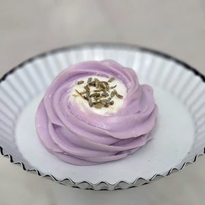 May include: A lavender-colored, swirl-shaped dessert topped with white cream and dried lavender buds, presented on a white, fluted, metal plate. The dessert is the focal point, with a soft, blurred background.