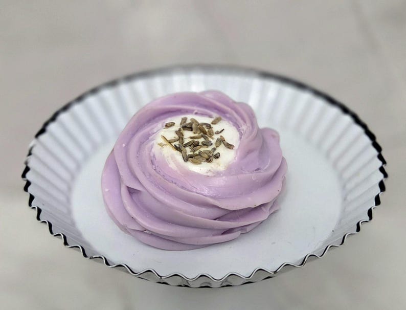 May include: A lavender-colored, swirl-shaped dessert topped with white cream and dried lavender buds, presented on a white, fluted, metal plate. The dessert is the focal point, with a soft, blurred background.