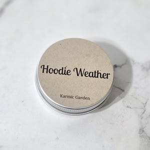 May include: A round metal tin with a light brown paper label on the lid. The label features the text "Hoodie Weather" in a dark, stylized script font, with the smaller text "Karmic Garden" printed in a simple sans-serif font below it.