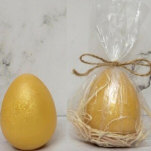 K&ouml;nnte beinhalten: Ein goldenes Ei mit schimmerndem Finish liegt neben einem Ei, das in durchsichtiges Cellophan gewickelt und mit Bindfaden verschn&uuml;rt ist. Das verpackte Ei liegt in einem Nest aus Stroh. Die Eier sind wahrscheinlich dekorativ oder f&uuml;r Ostern.