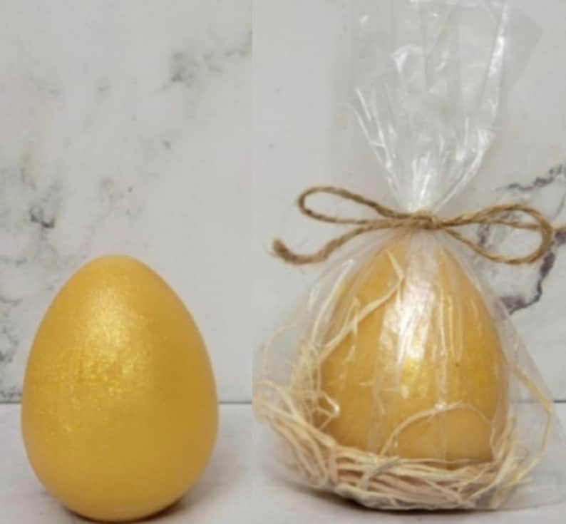 K&ouml;nnte beinhalten: Ein goldenes Ei mit schimmerndem Finish liegt neben einem Ei, das in durchsichtiges Cellophan gewickelt und mit Bindfaden verschn&uuml;rt ist. Das verpackte Ei liegt in einem Nest aus Stroh. Die Eier sind wahrscheinlich dekorativ oder f&uuml;r Ostern.