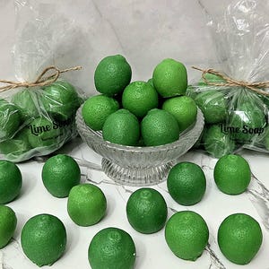 May include: Green lime-shaped soaps are displayed in a clear glass bowl and in clear cellophane bags tied with twine. The bags are labeled "Lime Soap". Several loose soaps are scattered around the bowl.