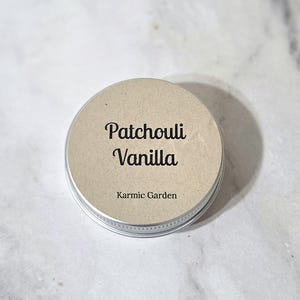 May include: A round metal tin with a beige paper label featuring the black text Patchouli Vanilla and Karmic Garden. The container has a silver-colored rim and a flat top, presenting a simple and functional design for a small cosmetic or balm product.