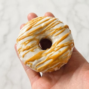 May include: A round donut with a smooth white glaze and a decorative drizzle of golden-brown caramel icing. The donut features a central hole and a circular shape, showcasing a contrast between the opaque white base and the glossy amber-colored topping.