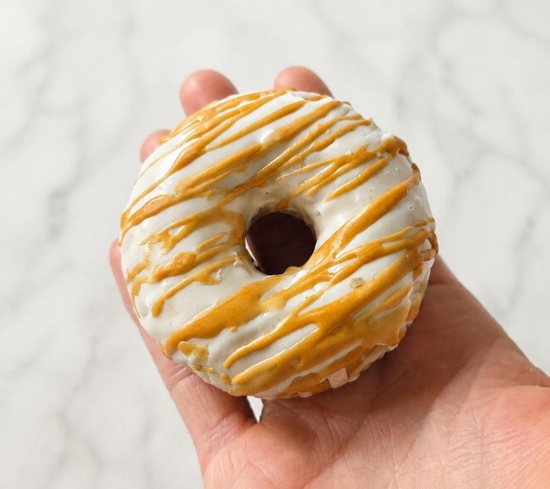 May include: A round donut with a smooth white glaze and a decorative drizzle of golden-brown caramel icing. The donut features a central hole and a circular shape, showcasing a contrast between the opaque white base and the glossy amber-colored topping.