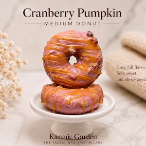May include: Two stacked cranberry pumpkin medium donut-shaped bars with pink glaze and caramel-colored drizzle on a white pedestal. Text reads: Cranberry Pumpkin, MEDIUM DONUT, Cozy fall flavor. Soft, sweet, and oh-so-good. Karmic Garden THE FACIAL BAR APOTHECARY.