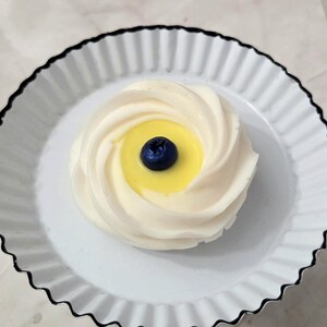 May include: A single dessert, resembling a meringue, sits on a white plate with a black scalloped edge. The dessert has a swirl design with a yellow center and a single blueberry on top.