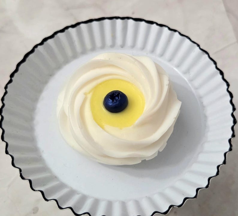 May include: A single dessert, resembling a meringue, sits on a white plate with a black scalloped edge. The dessert has a swirl design with a yellow center and a single blueberry on top.