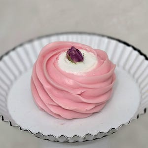 May include: A pink dessert with a swirl design, topped with a small purple flower, sits in a white, fluted dish with a silver rim. The dessert has a white center and is presented on a white surface.