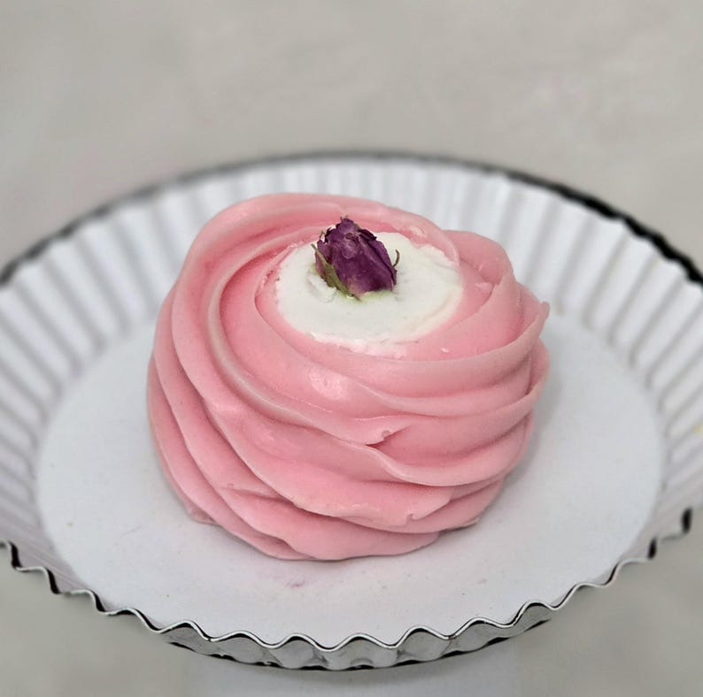 May include: A pink dessert with a swirl design, topped with a small purple flower, sits in a white, fluted dish with a silver rim. The dessert has a white center and is presented on a white surface.